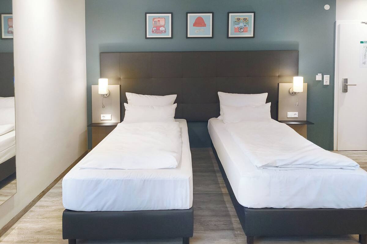 Habitación de hotel con dos camas individuales, cabecera oscura, mesitas de noche fijadas a la pared con lámparas, y cuadros enmarcados en la pared.