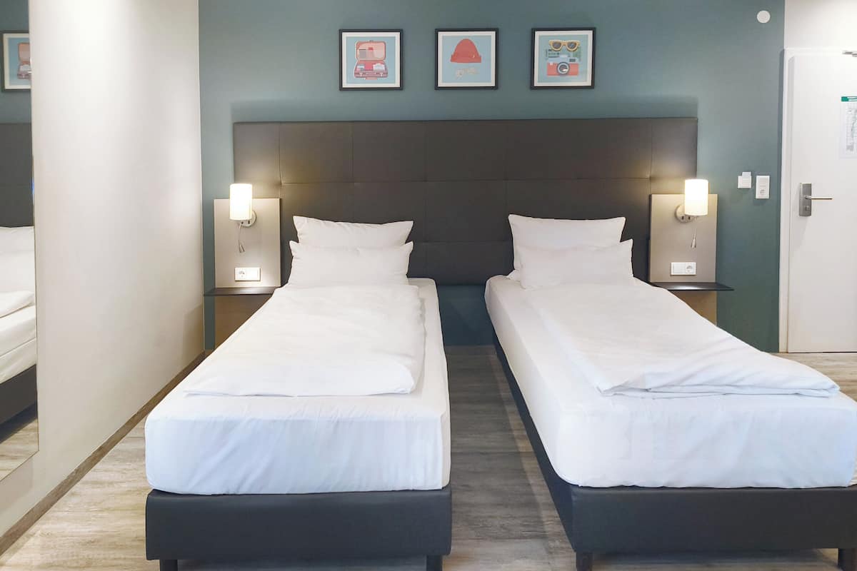 Quarto de hotel com duas camas de solteiro, cabeceira escura, mesas de cabeceira fixadas na parede com luminárias e quadros na parede.