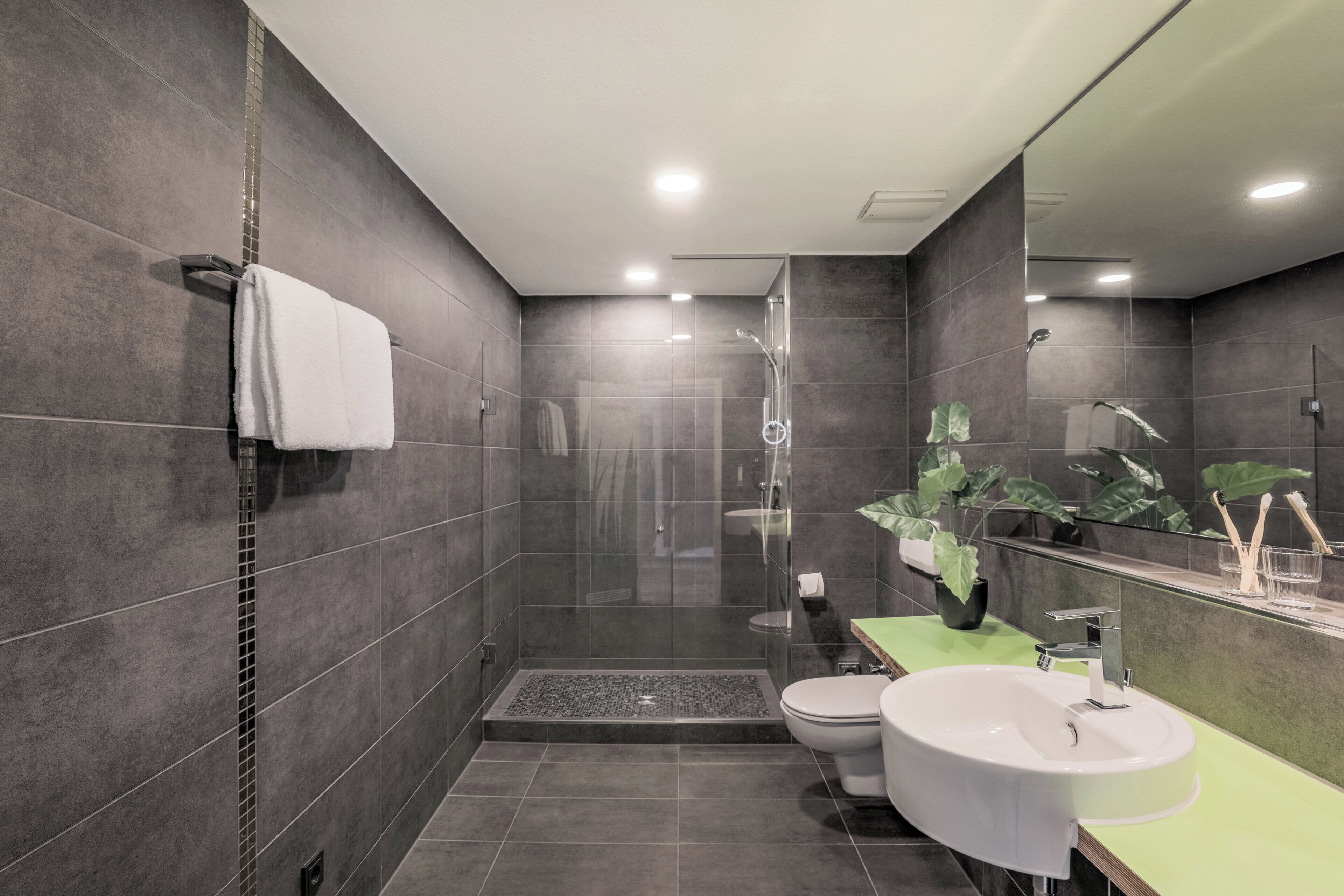 A modern bathroom with a shower, toilet, and sink, featuring dark tiles and green countertops.