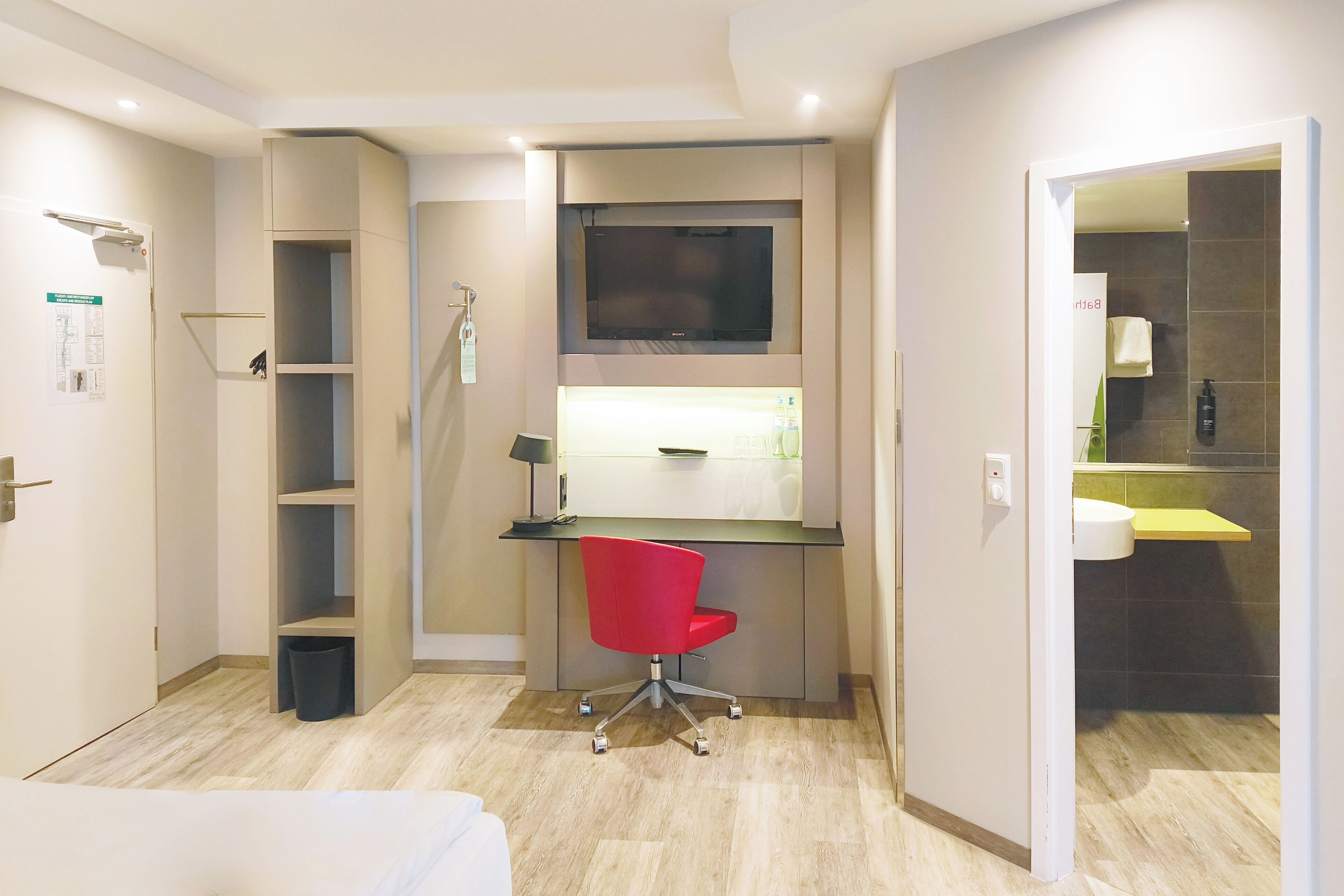 Een moderne hotelkamer met een bureau, een rode stoel, een televisie en een badkamer zichtbaar door een geopende deur.