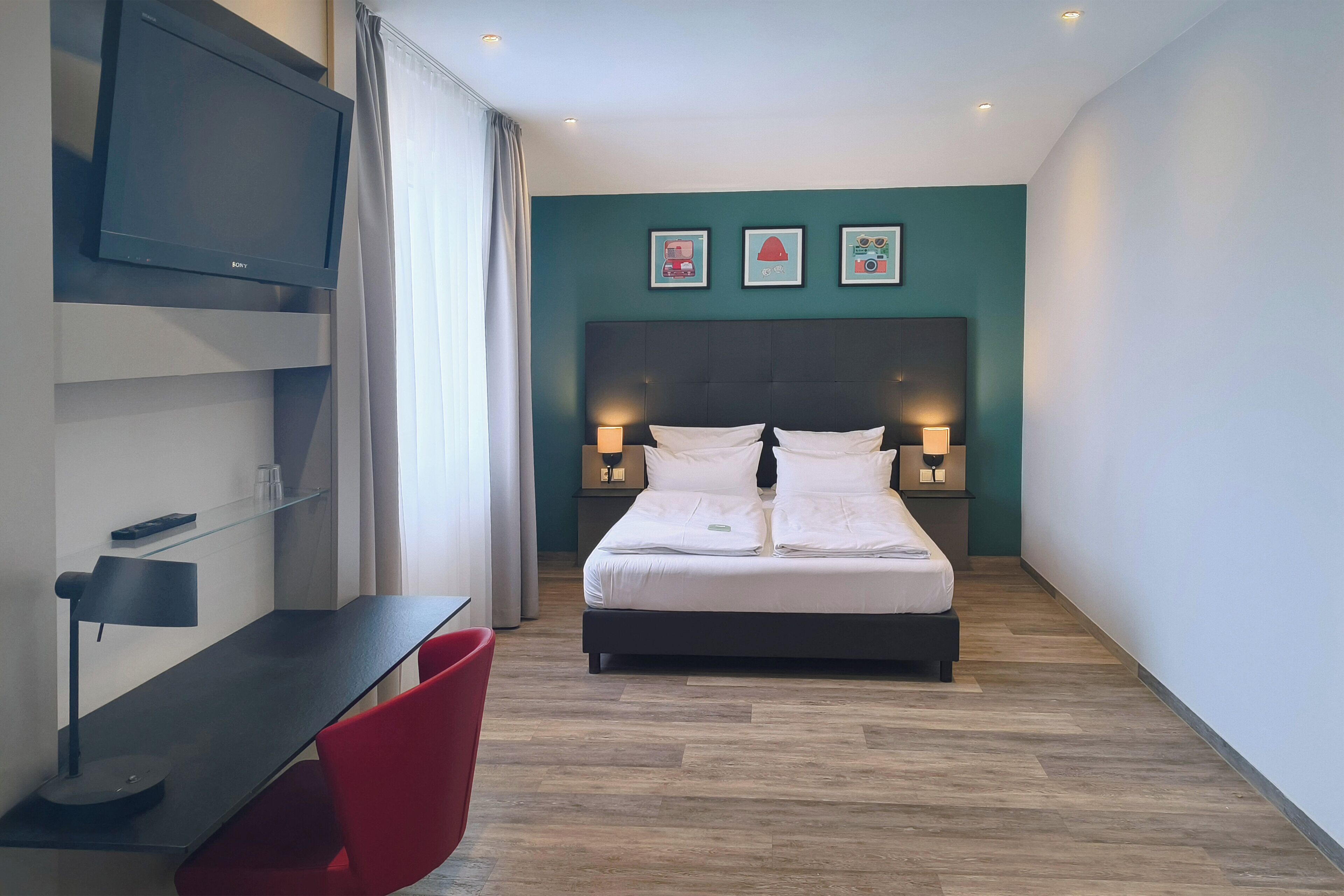 Quarto de hotel moderno com cama, abajures, televisão, escrivaninha com luminária e uma cadeira vermelha.