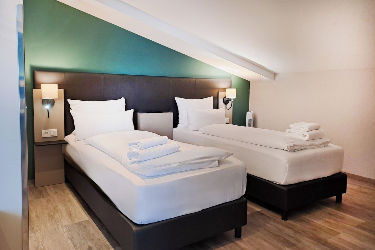 A hotel room with two beds, a green accent wall, and bedside lamps.