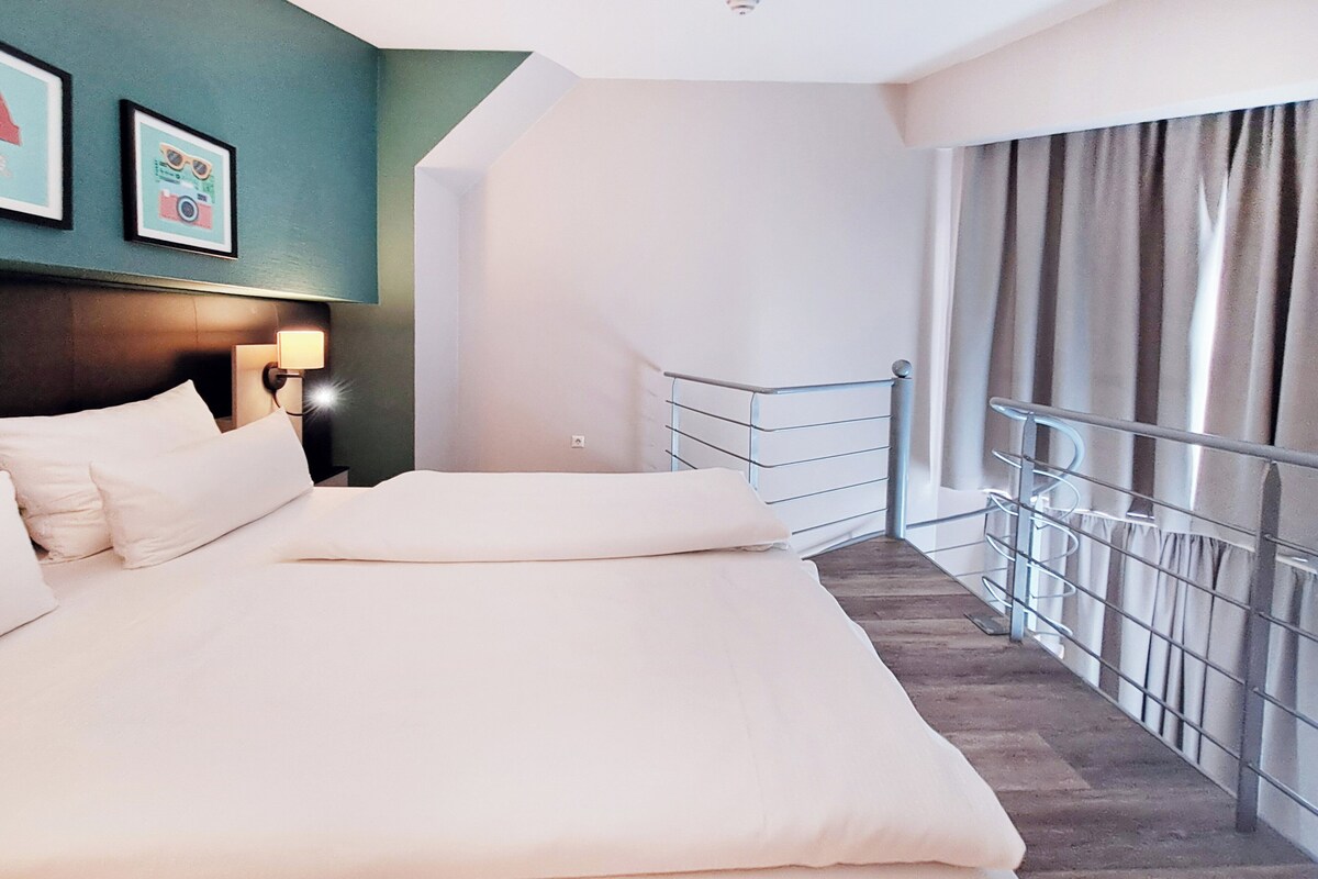 A hotel room with a large bed, a headboard with a light, a staircase with a railing, and a view of the outside.