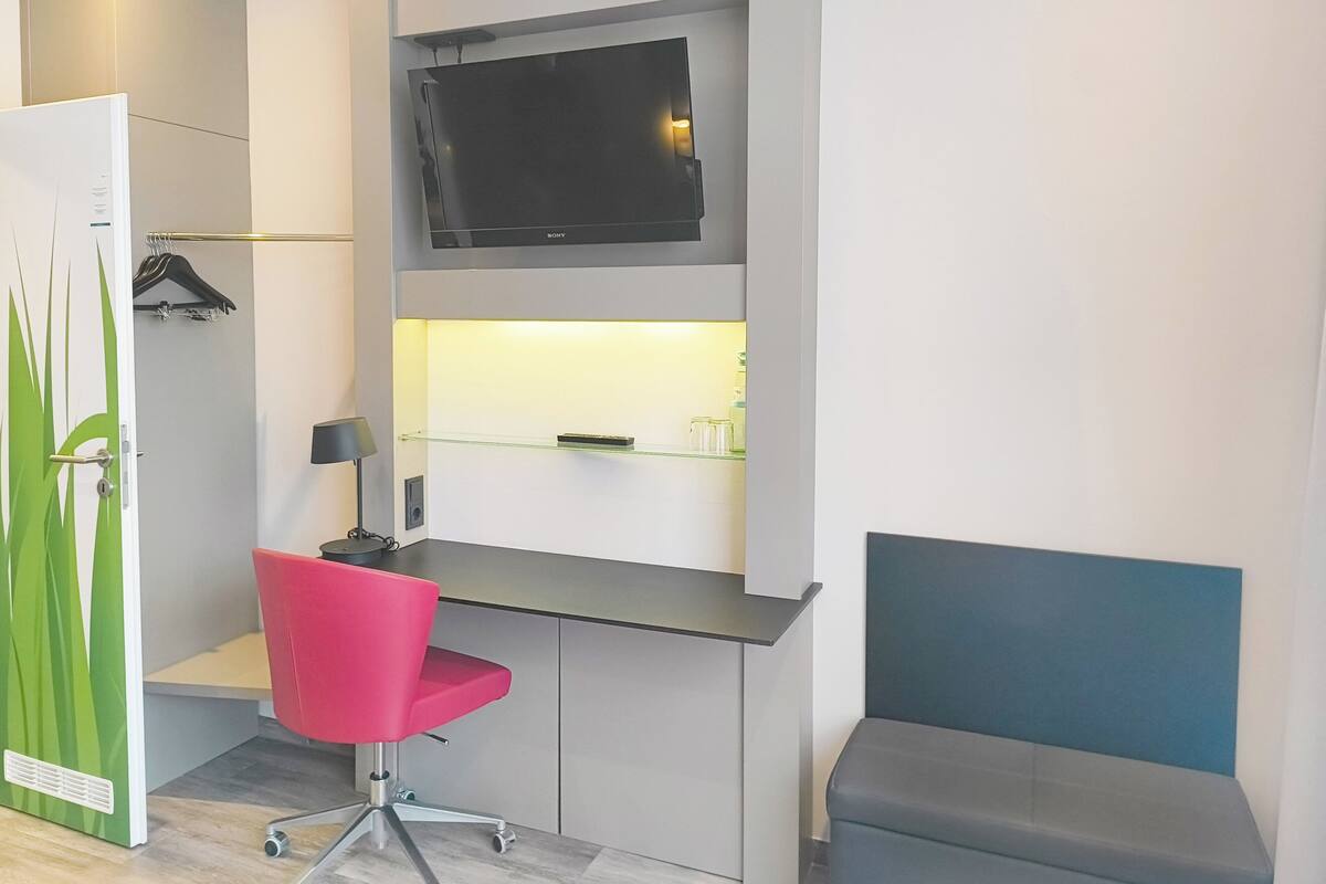 Una habitación moderna con escritorio, televisor de pantalla plana, una silla roja, un banco y una puerta verde.