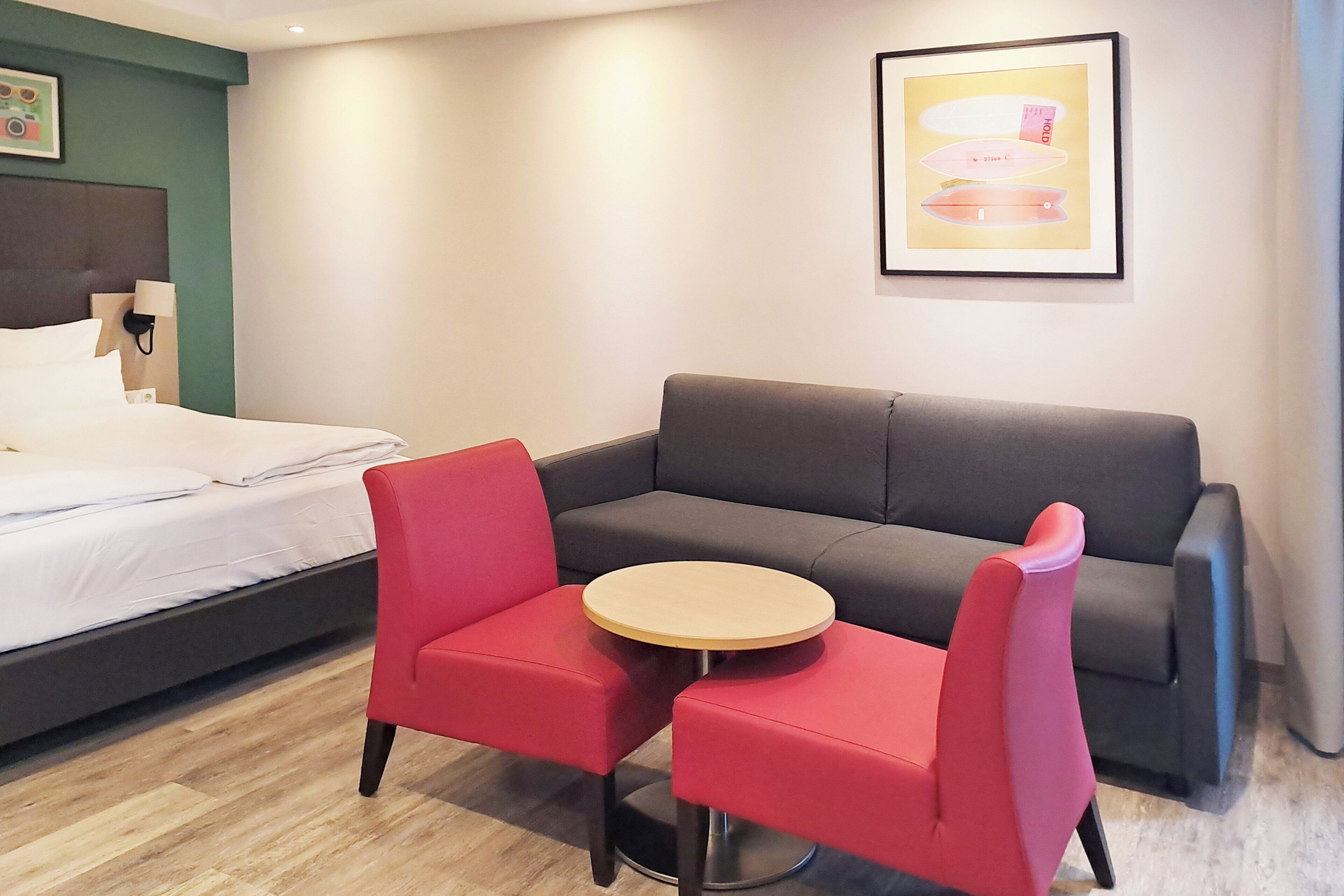 Ett hotellrum med en säng, en grå soffa, två röda stolar runt ett litet träbord och en inramad tavla på väggen.