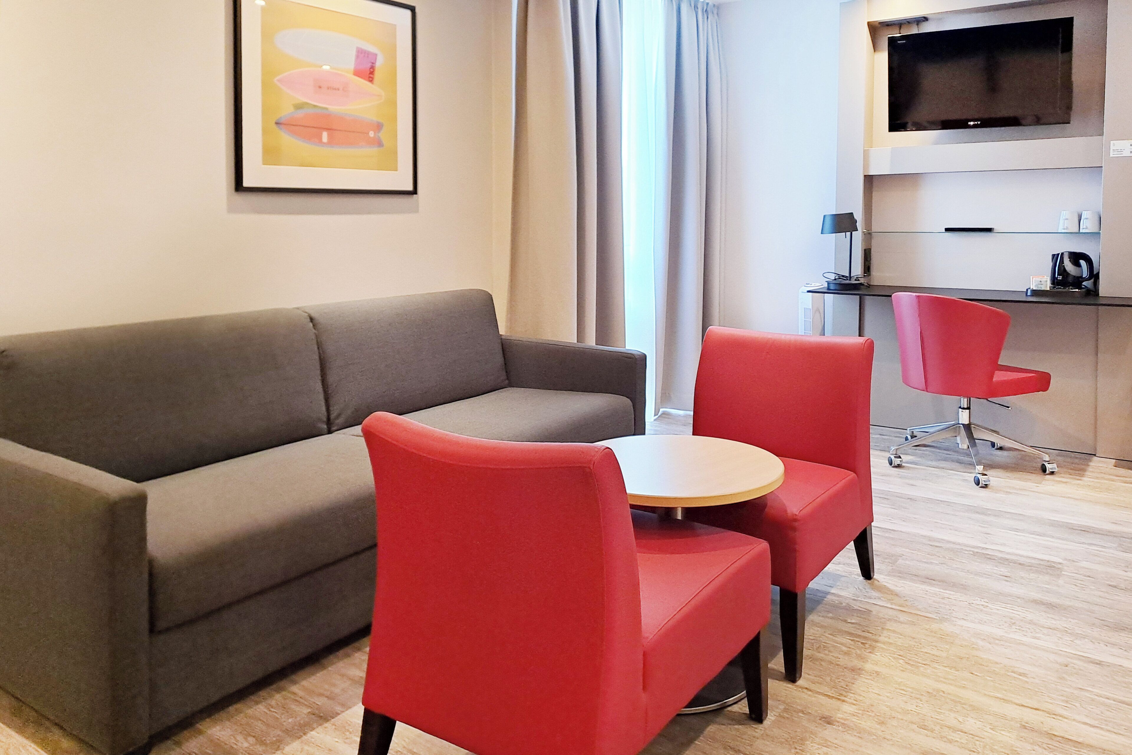 A modern living area with a grey sofa, a round table, and red chairs. A flat-screen TV is mounted on the wall above a kitchen counter.
