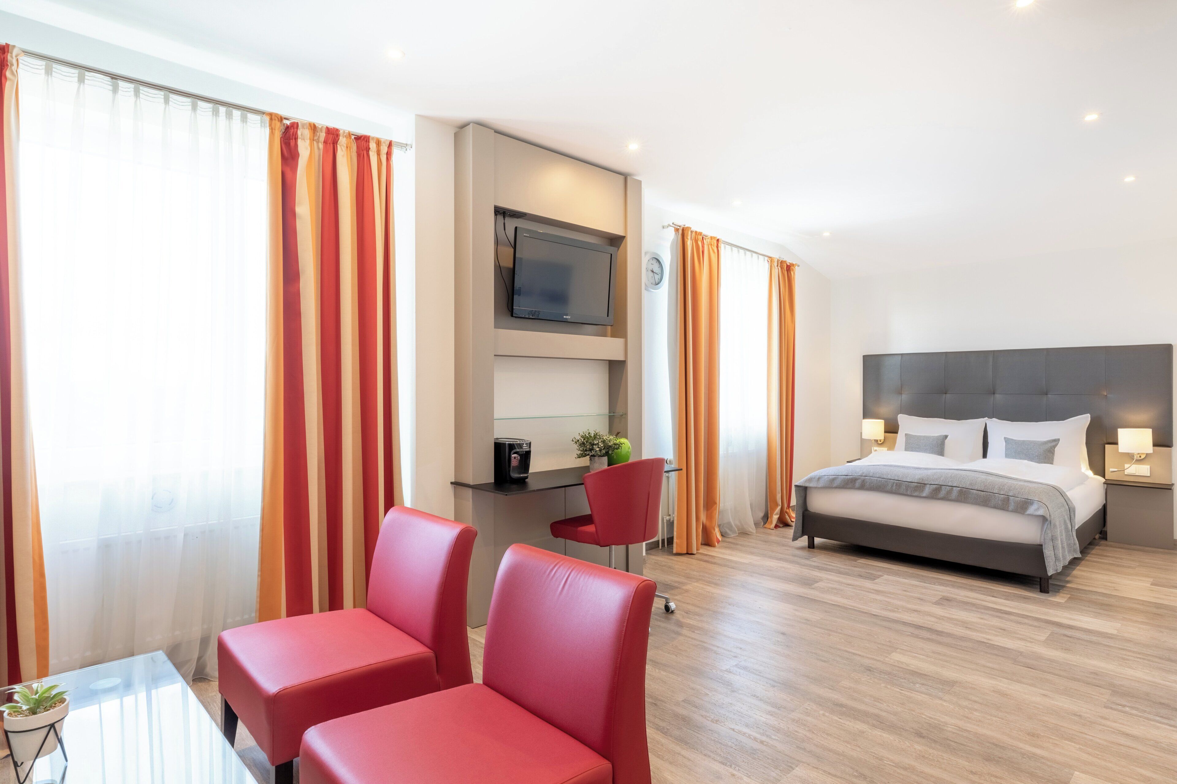 A hotel room with a bed, a desk, a TV, and two red chairs.