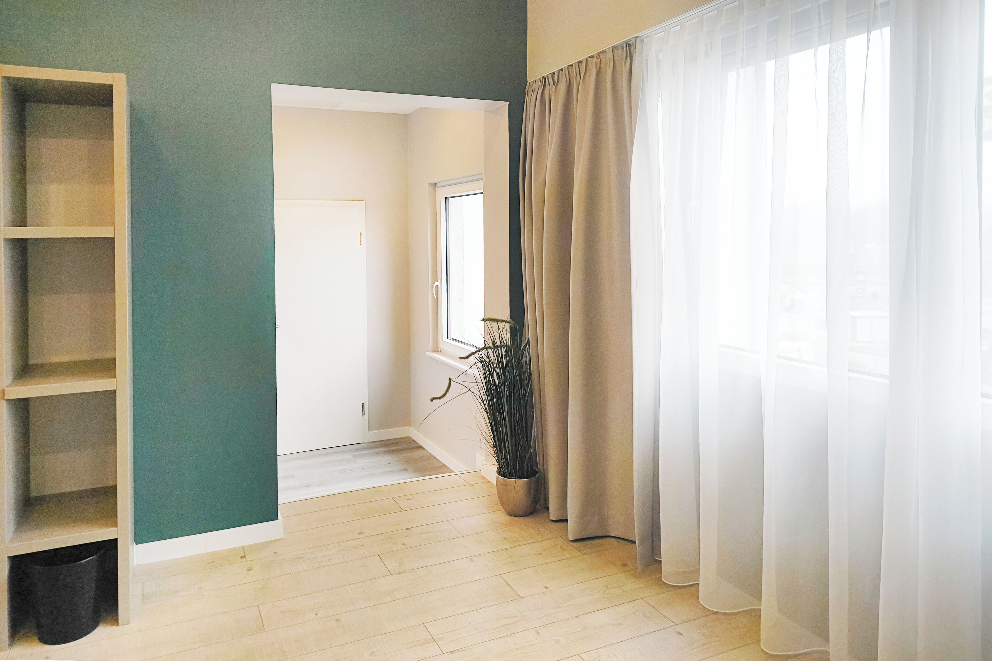 Una habitación con piso de madera, pared verde, puerta blanca, ventana con planta y cortinas de encaje.