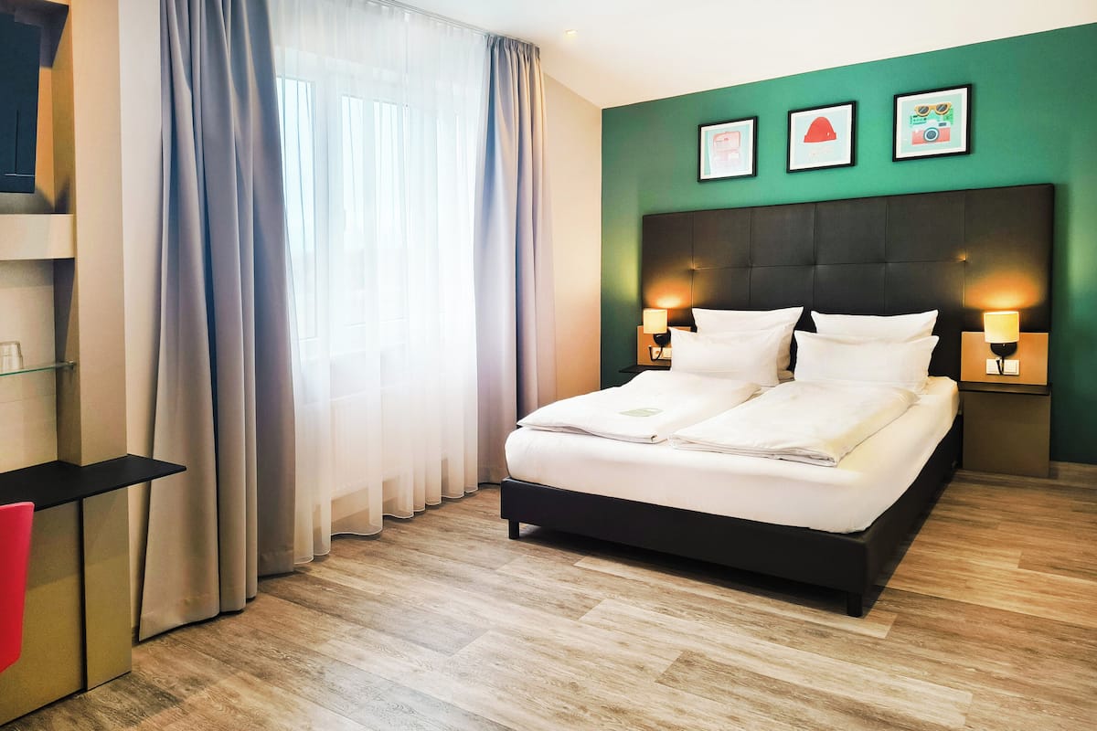 Quarto de hotel moderno com uma cama grande, luminárias de cabeceira, uma escrivaninha e vista para o exterior através de cortinas translúcidas.
