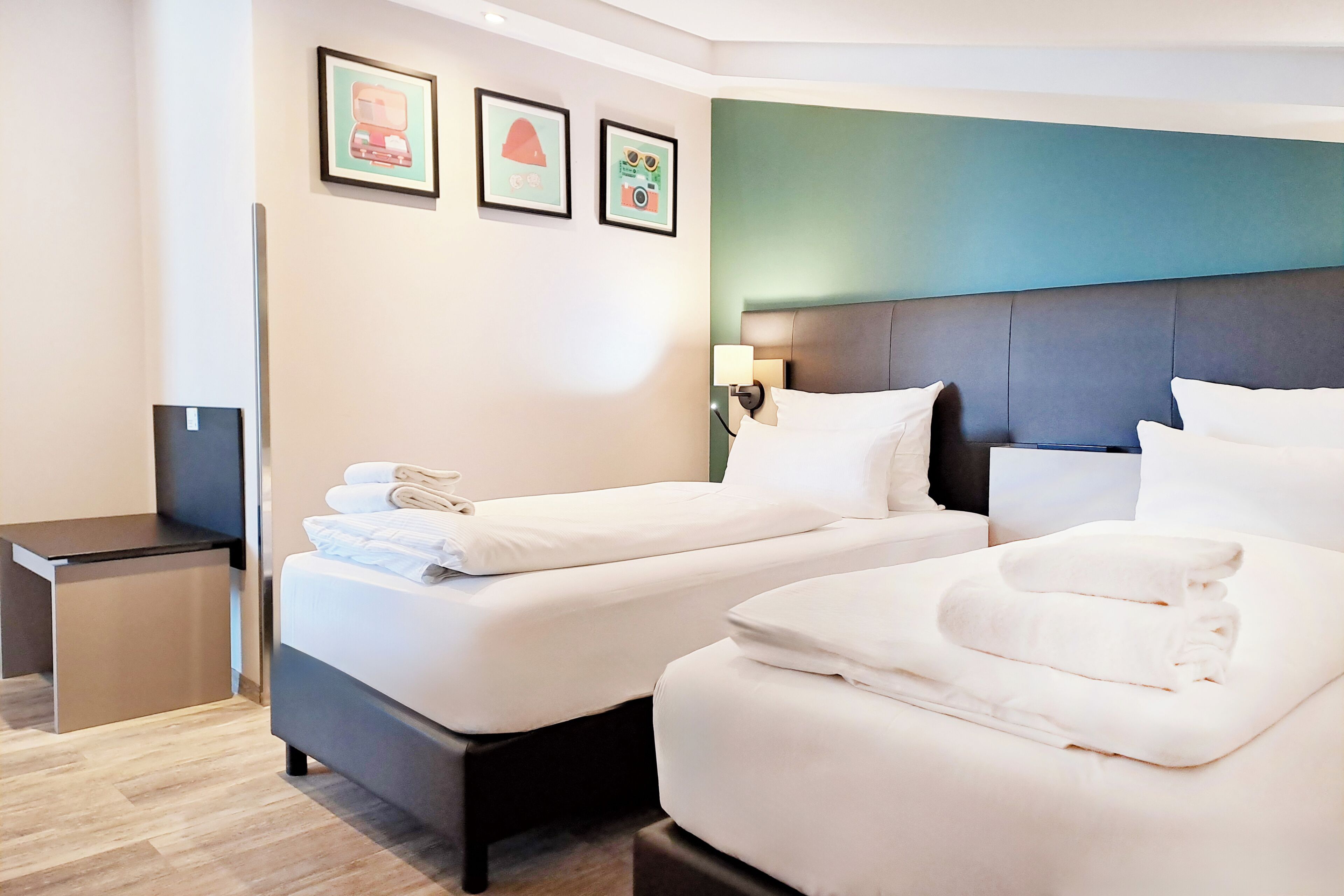 A hotel room with two beds, a small desk, and a wall with framed pictures.