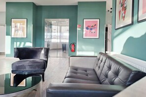 Un moderno vestíbulo con un sofá y sillón de cuero negro, una mesa de centro de vidrio, paredes verde azulado y obras de arte enmarcadas.