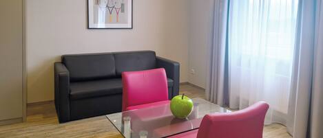A modern living room with a glass-top table, a green apple, a black sofa, and a pink chair.