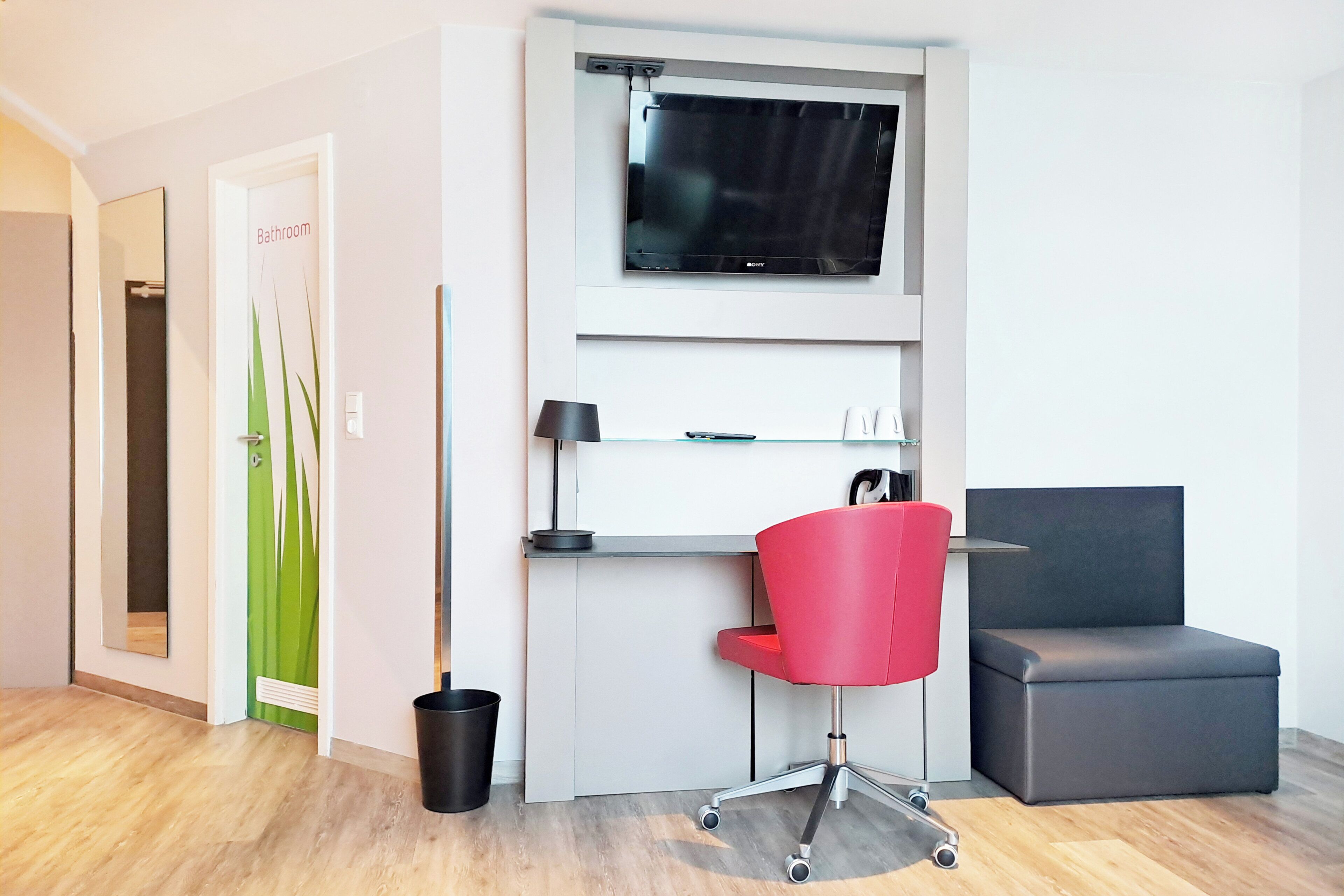 Une chambre moderne équipée d’un téléviseur à écran plat, d’un fauteuil de bureau rouge, d’un canapé gris et d’un petit bureau avec une lampe.