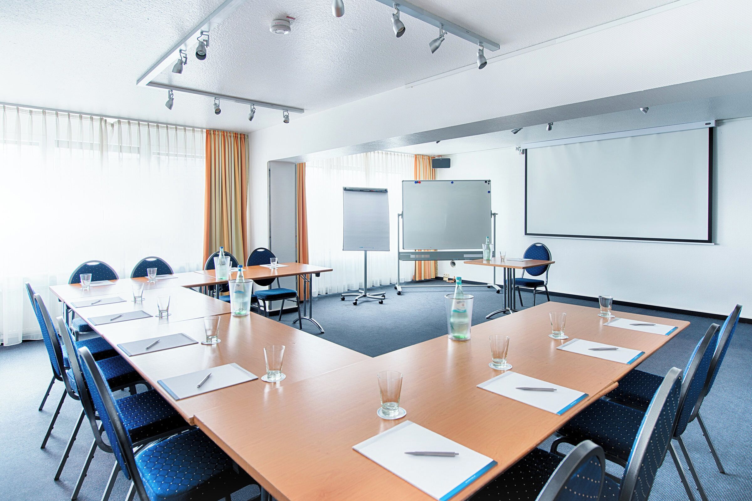 Ein Konferenzraum mit langem Tisch, Stühlen, einer Whiteboard und einer Projektionsleinwand.