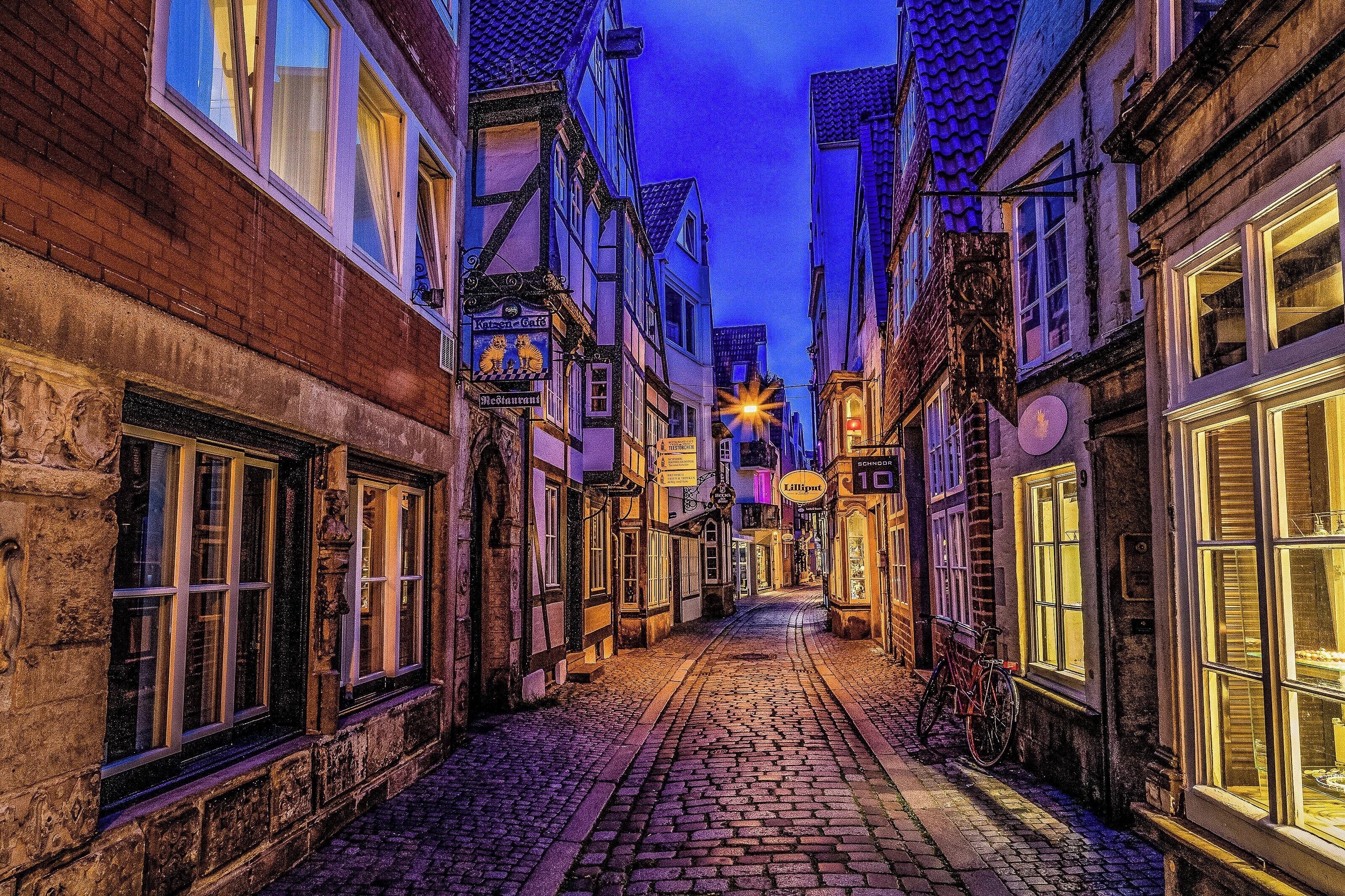 Una estrecha calle adoquinada flanqueada por edificios tradicionales europeos, iluminada por farolas.