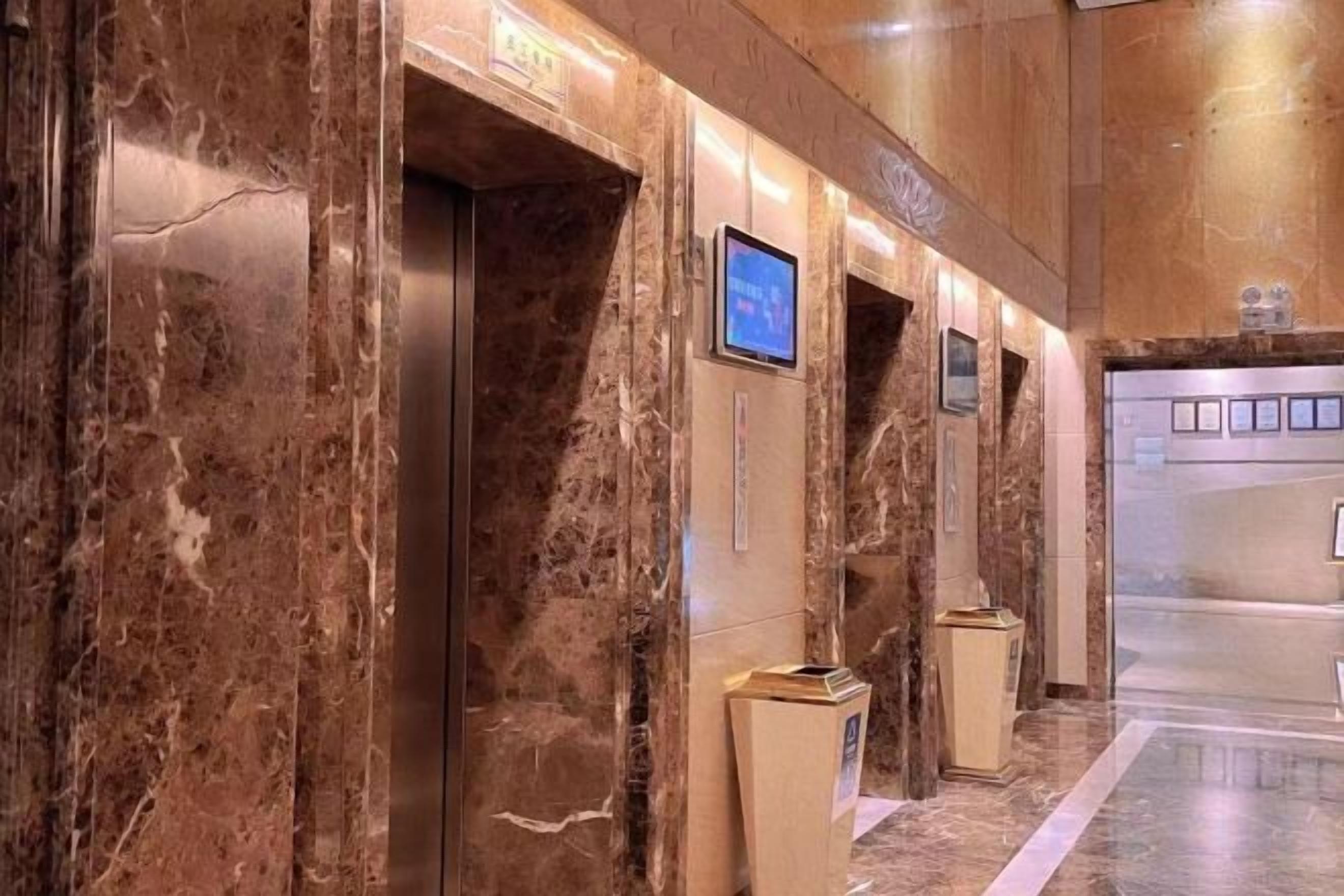 Zona de ascensores con paredes de mármol, una pantalla informativa digital y un mostrador de recepción.