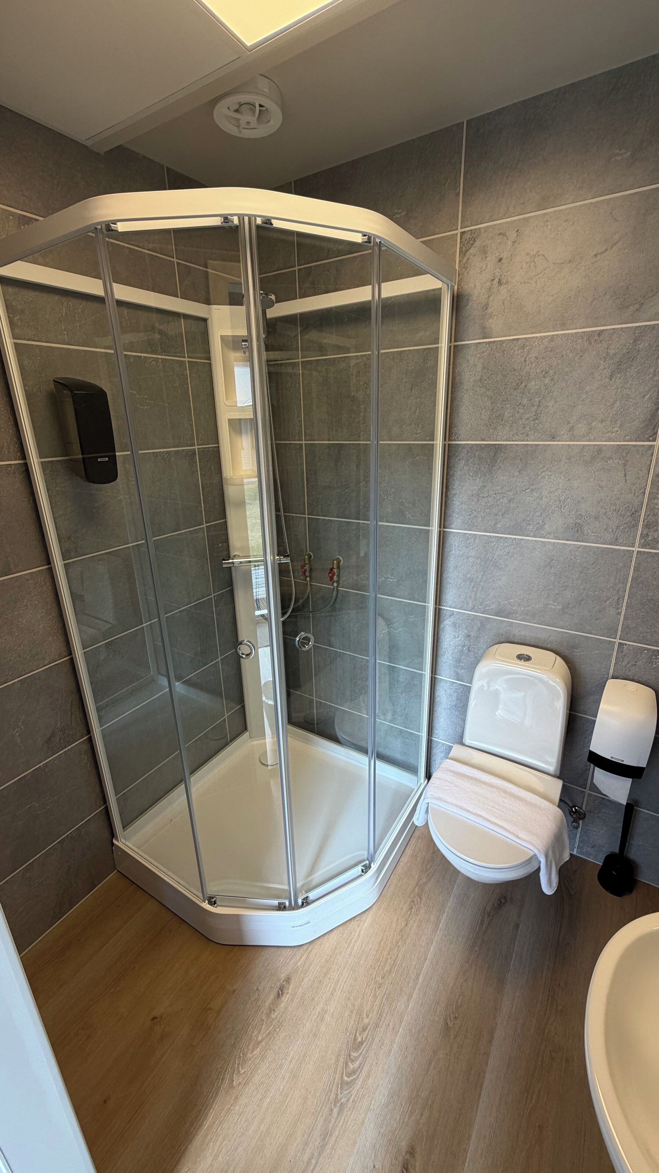 Douchecabine met hoekdouche, glazen deur, toilet met handdoek en desinfecterende handpompen.
