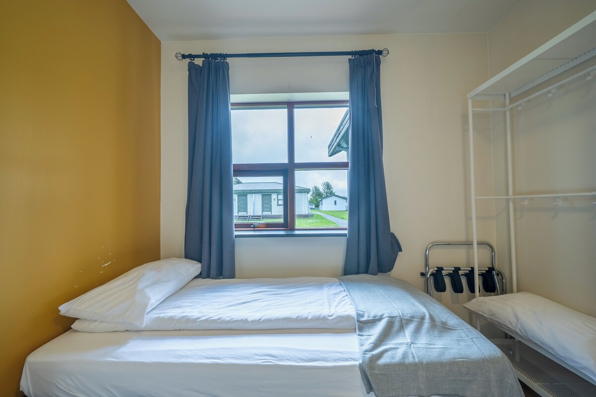 Uma cama de solteiro com roupa de cama branca, uma janela com cortinas azuis e uma estante de metal com objetos pretos.