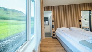 Een slaapkamer met een groot raam, een wand van houten panelen, een bed, een nachtkastje en een spiegel.
