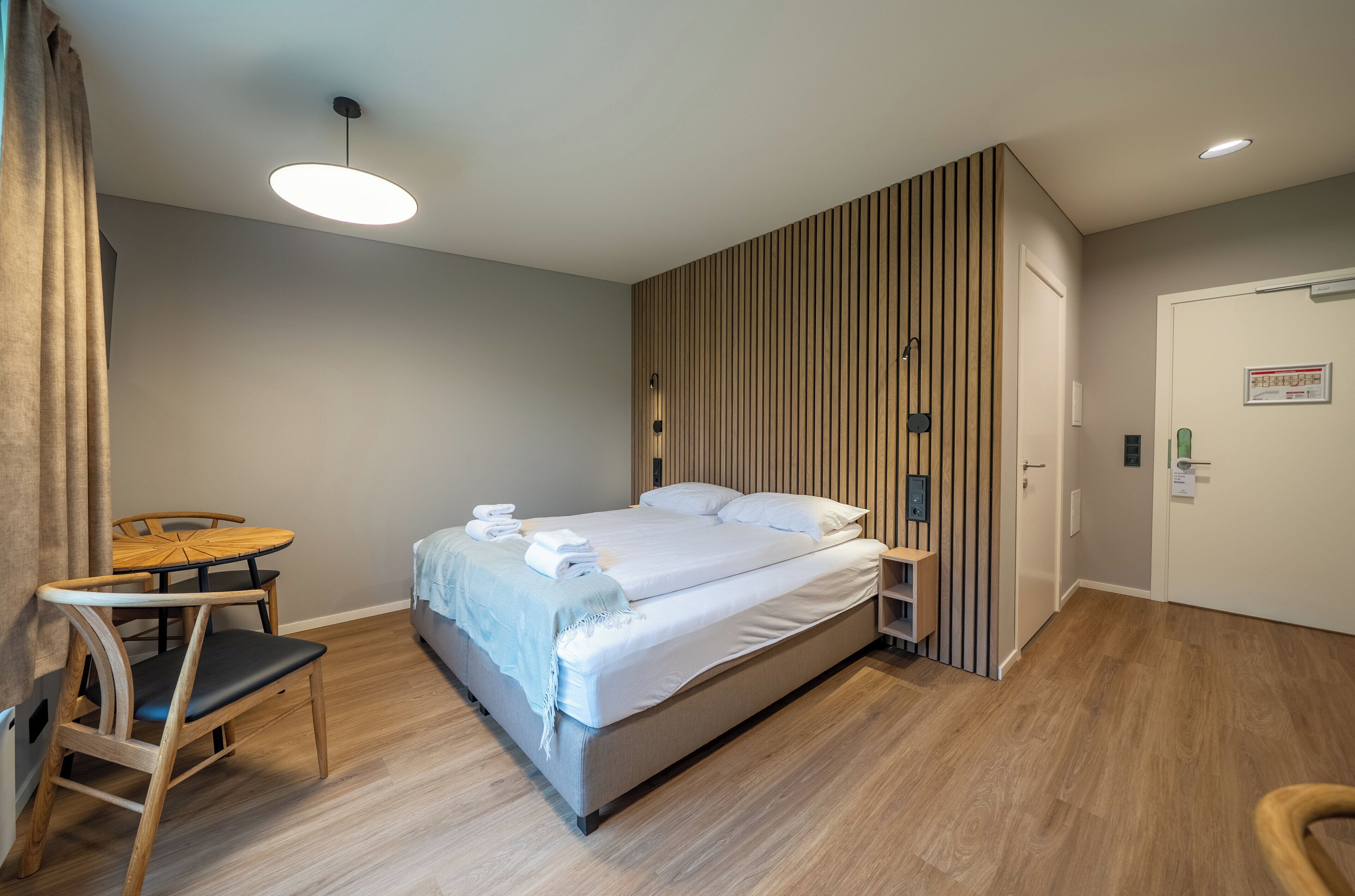Una habitación de hotel moderna con una cama, una silla de madera, una mesita pequeña y un estante en la pared.