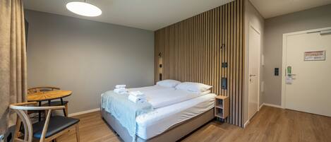 Una habitación de hotel moderna con una cama, una silla de madera, una mesita pequeña y un estante en la pared.