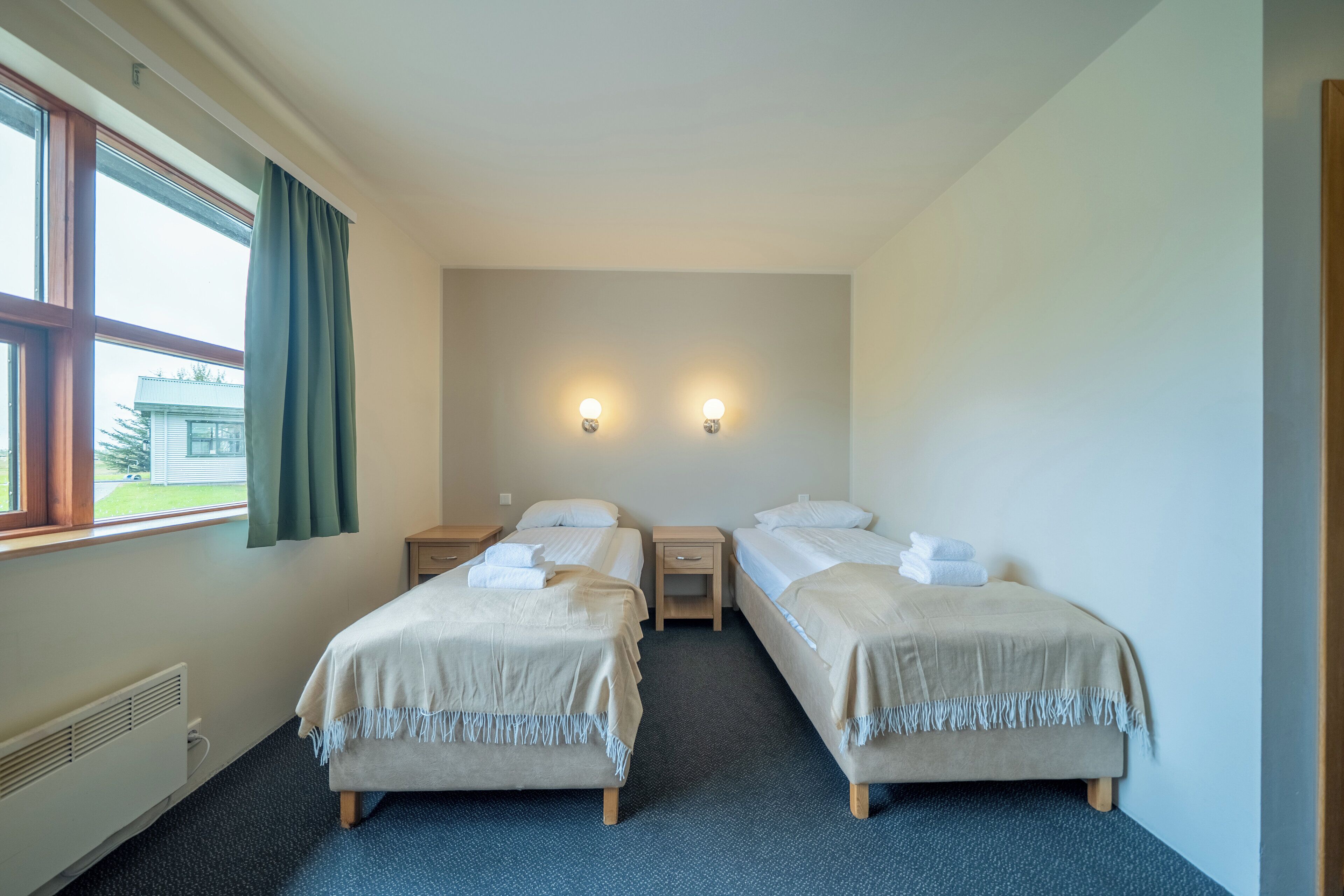 A hotel room with two single beds, a small bedside table, a nightstand, a window with curtains, and two wall-mounted lights.