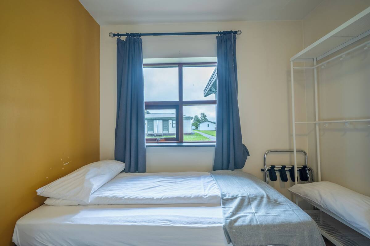 Una cama individual con ropa de cama blanca, una ventana con cortinas azules y una estantería metálica con objetos negros.