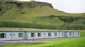 Een rij gelijkvloerse, metalen vakantiewoningen met groen gekleurde ramen, gelegen voor een grasachtige heuvel en bebost gebied.