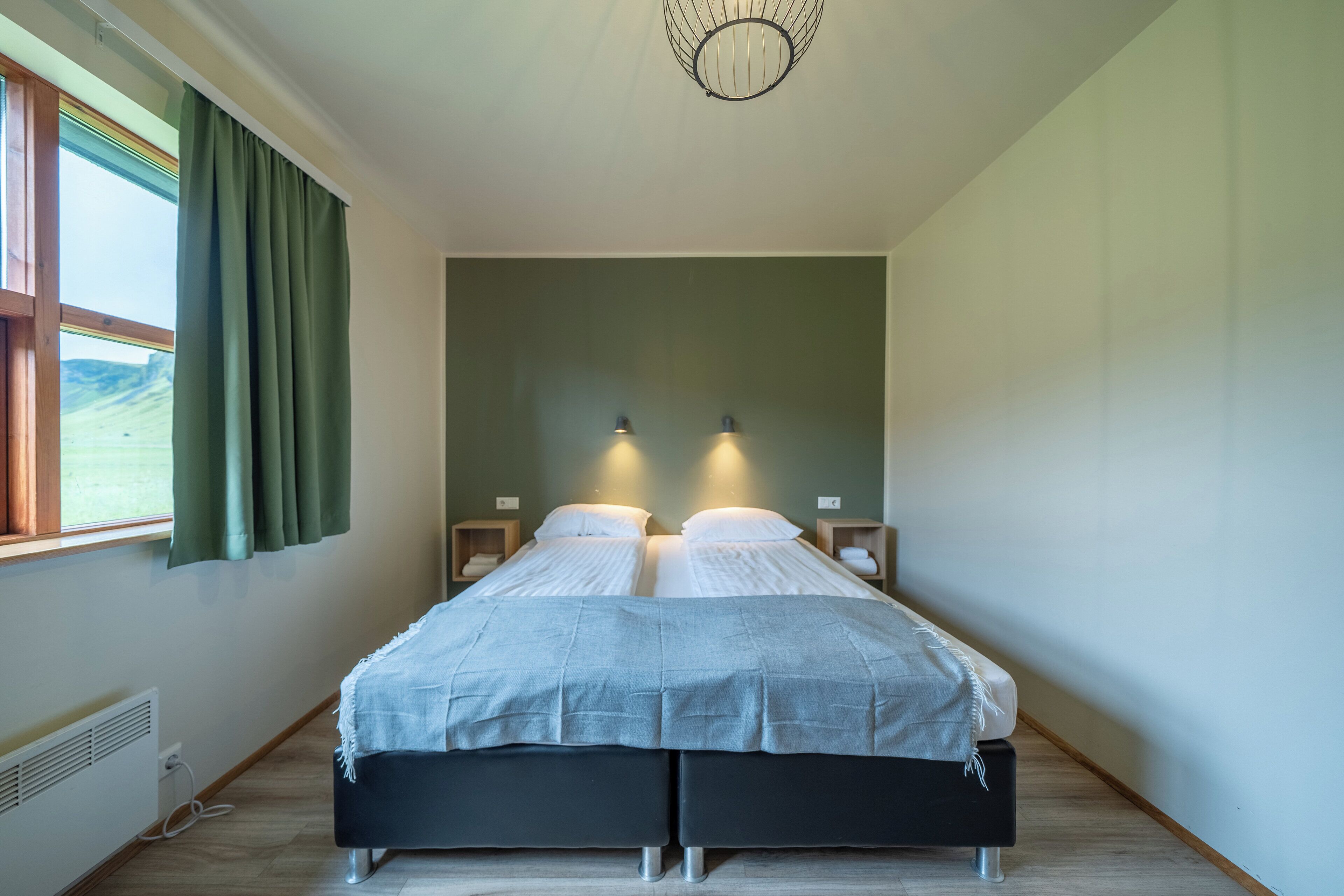 Ein Doppelbett mit grauer Bettdecke, zwei Nachttische mit Lampen, ein Fenster mit grünen Vorhängen und eine wandmontierte Leuchte.