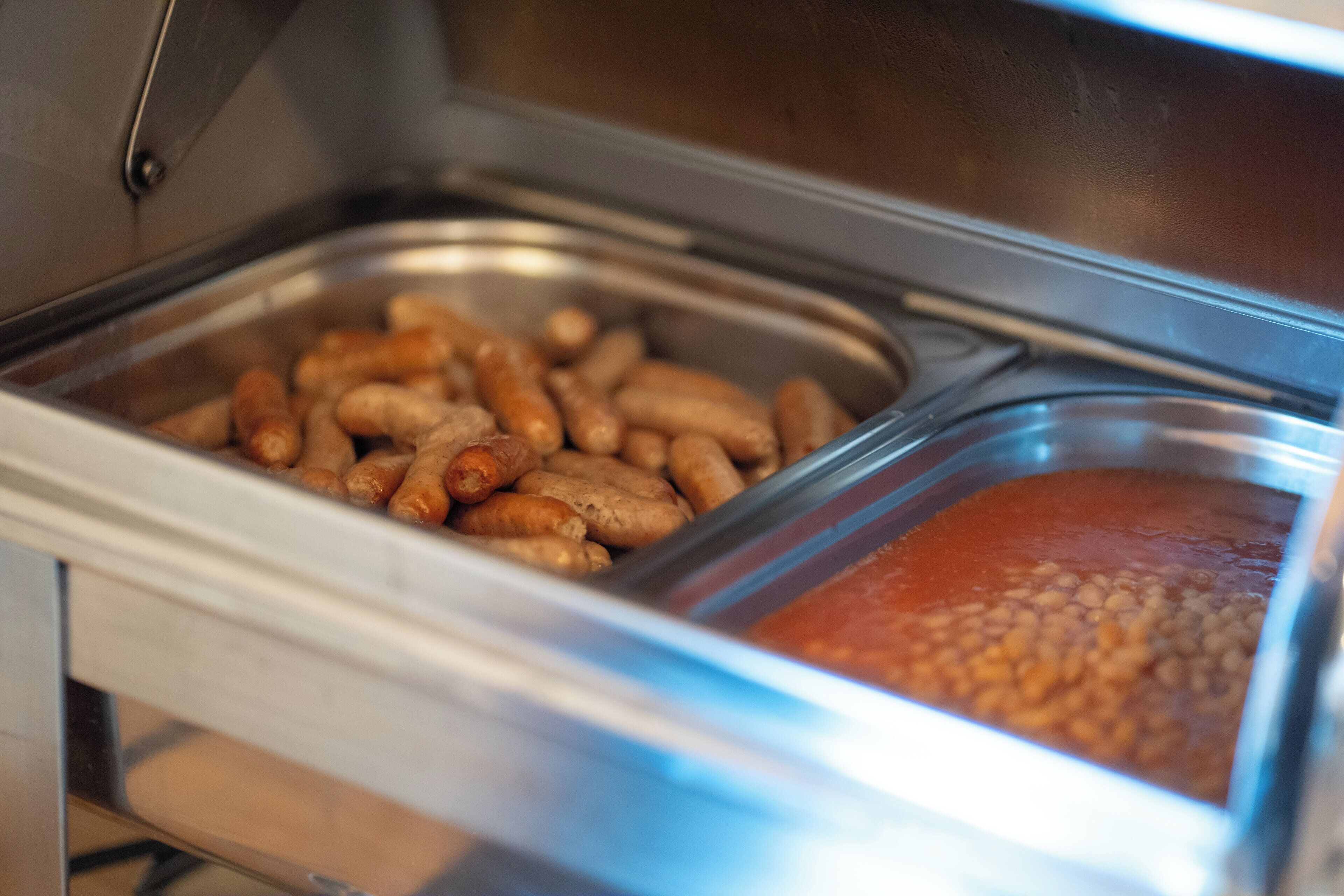A buffet with two metal trays, one containing sausages and the other with beans.