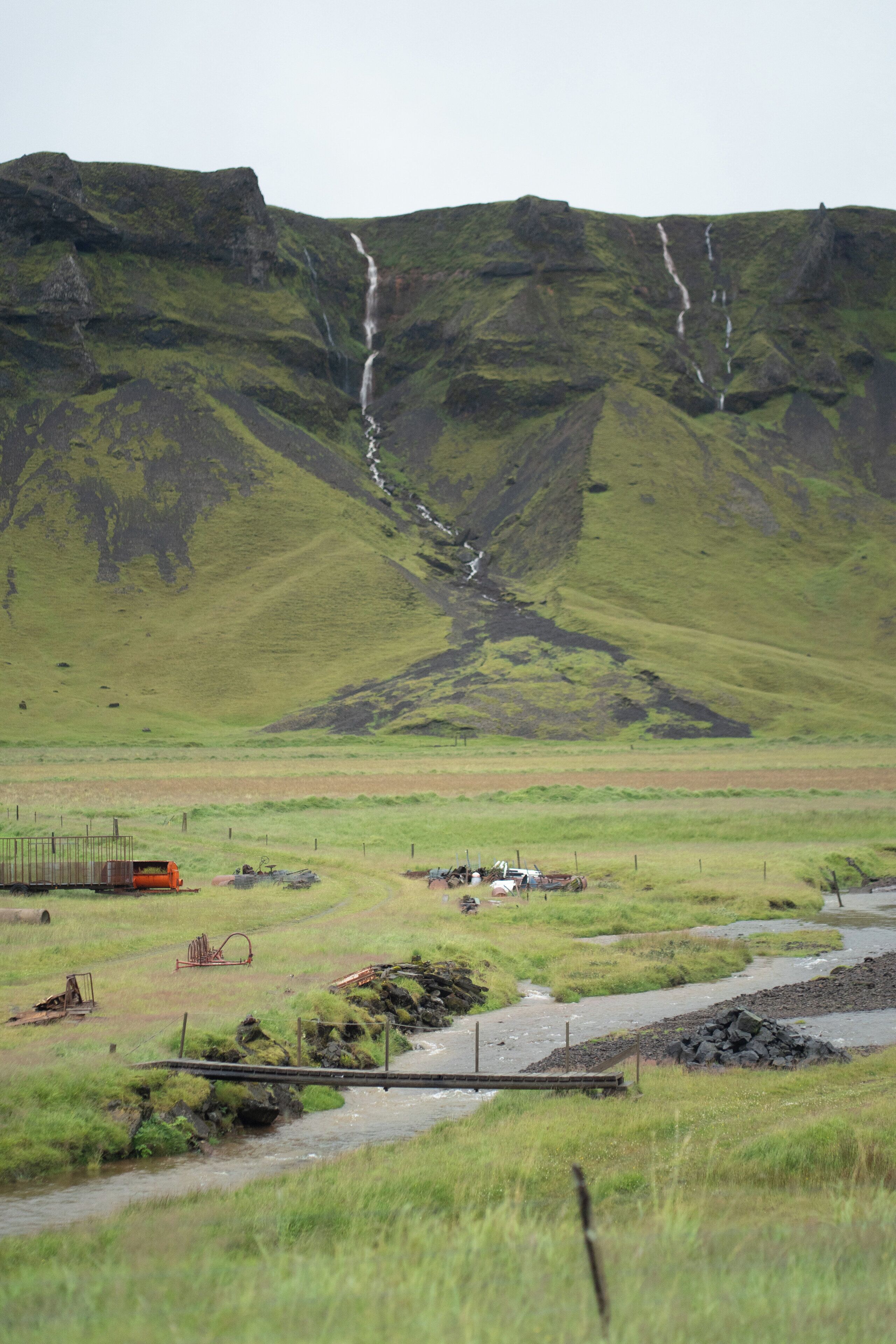 A rural landscape with a river, a small bridge, and a distant waterfall.
