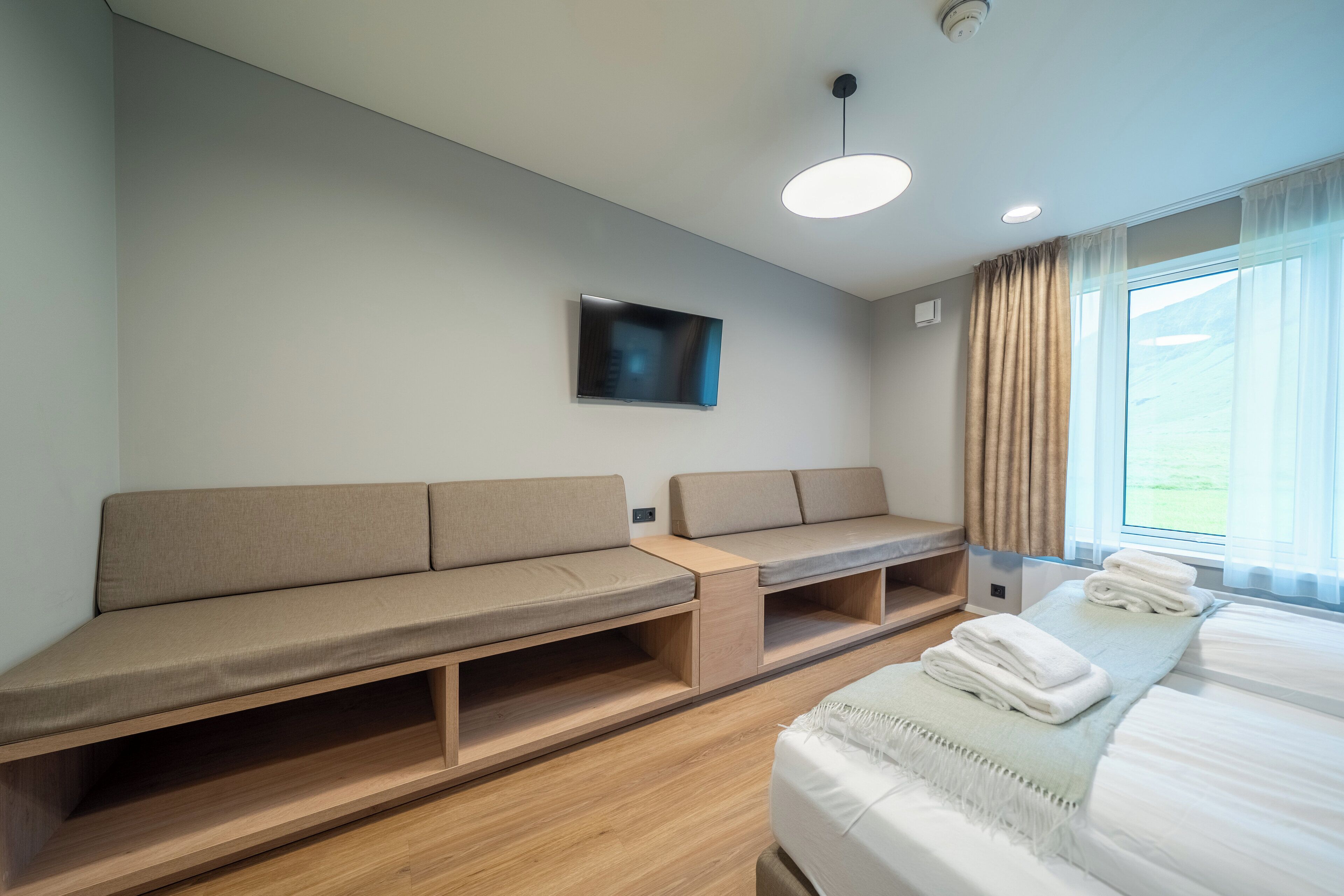 Moderni hotellihuone, jossa on sohva, seinälle asennettu televisio, valkoisella peitteellä varustettu sänky ja ikkuna, jossa on verhot.