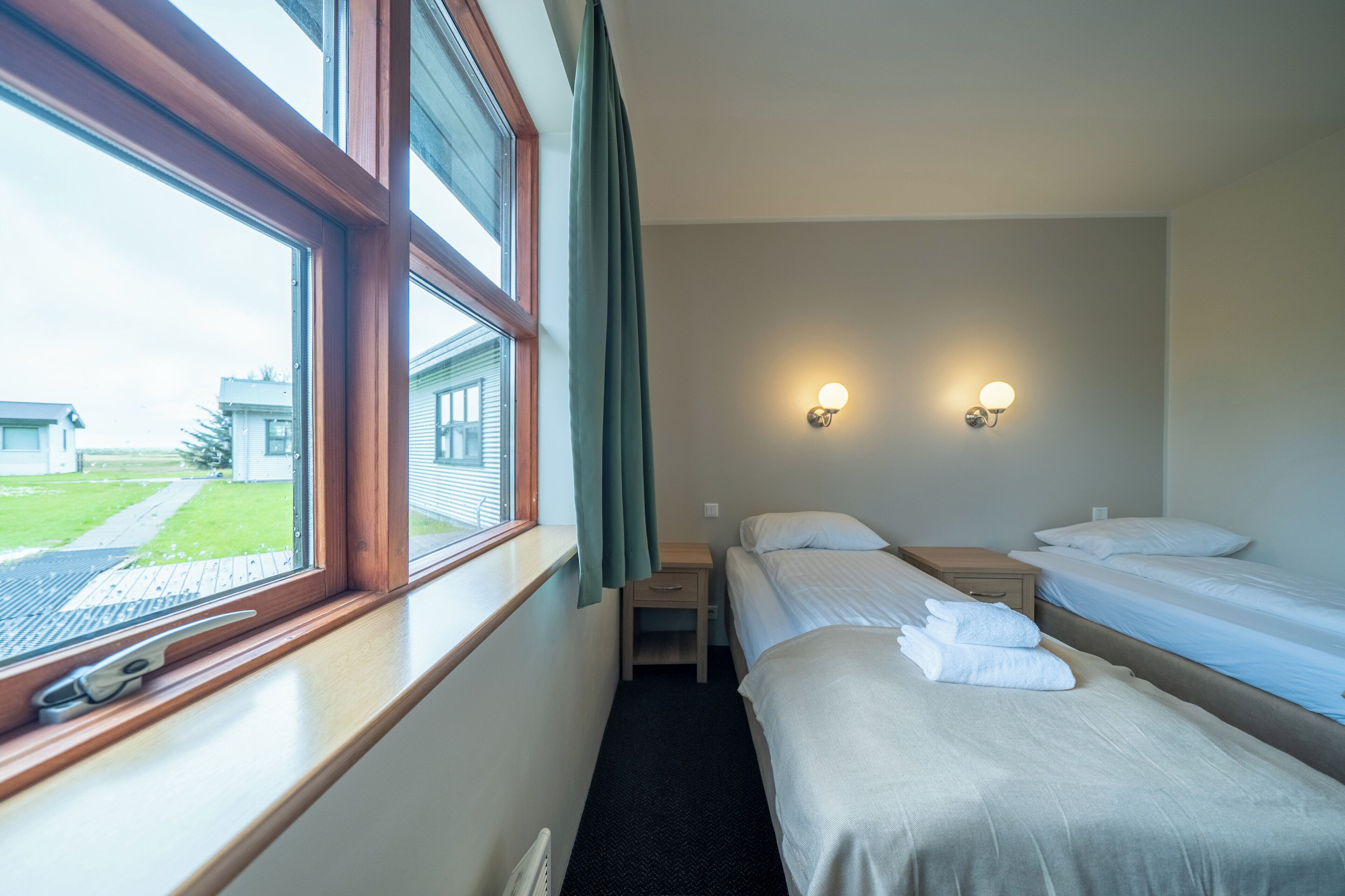 Habitación de hotel con dos camas individuales, una mesita de noche pequeña, un velador, una ventana con cortinas y dos luces fijadas a la pared.