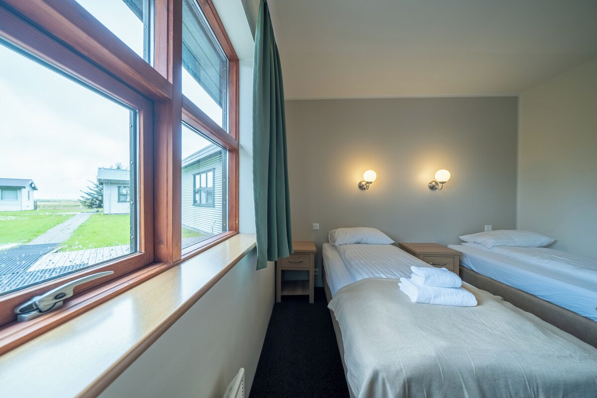 Quarto de hotel com duas camas de solteiro, uma pequena mesa de cabeceira, um criado-mudo, uma janela com cortinas e duas luminárias fixadas na parede.