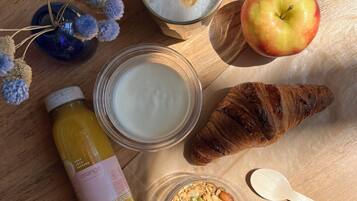 Un desayuno con un croissant, una manzana, yogur, granola y una botella de jugo sobre una mesa de madera.