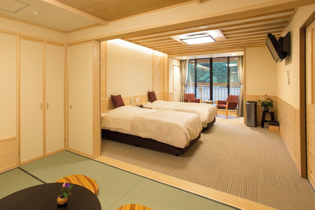 A hotel room with two beds, a small table with a vase, and a view of the outdoors through large windows.