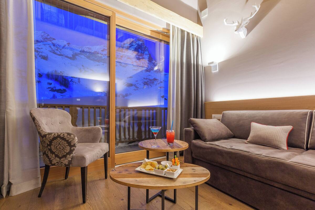 A cozy room with a sofa, armchair, and a small table with snacks and a drink. A large window with a view of a snowy landscape is visible.