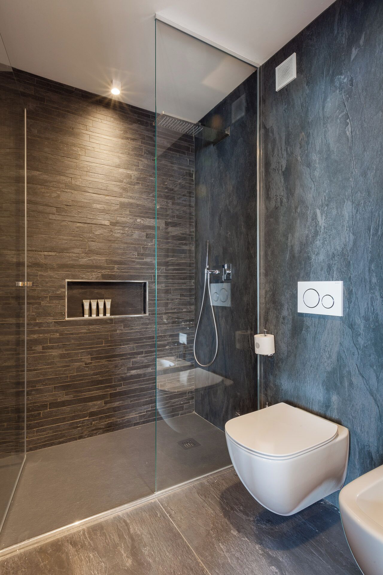 A modern bathroom with a glass-enclosed shower, a built-in shelf with toiletries, and a toilet with a bidet attachment.