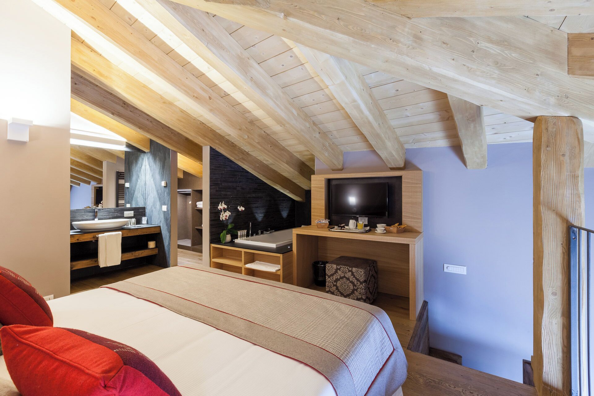 A modern bedroom with a sloped ceiling, a large bed, a TV, and a bathroom with a large mirror and sink.