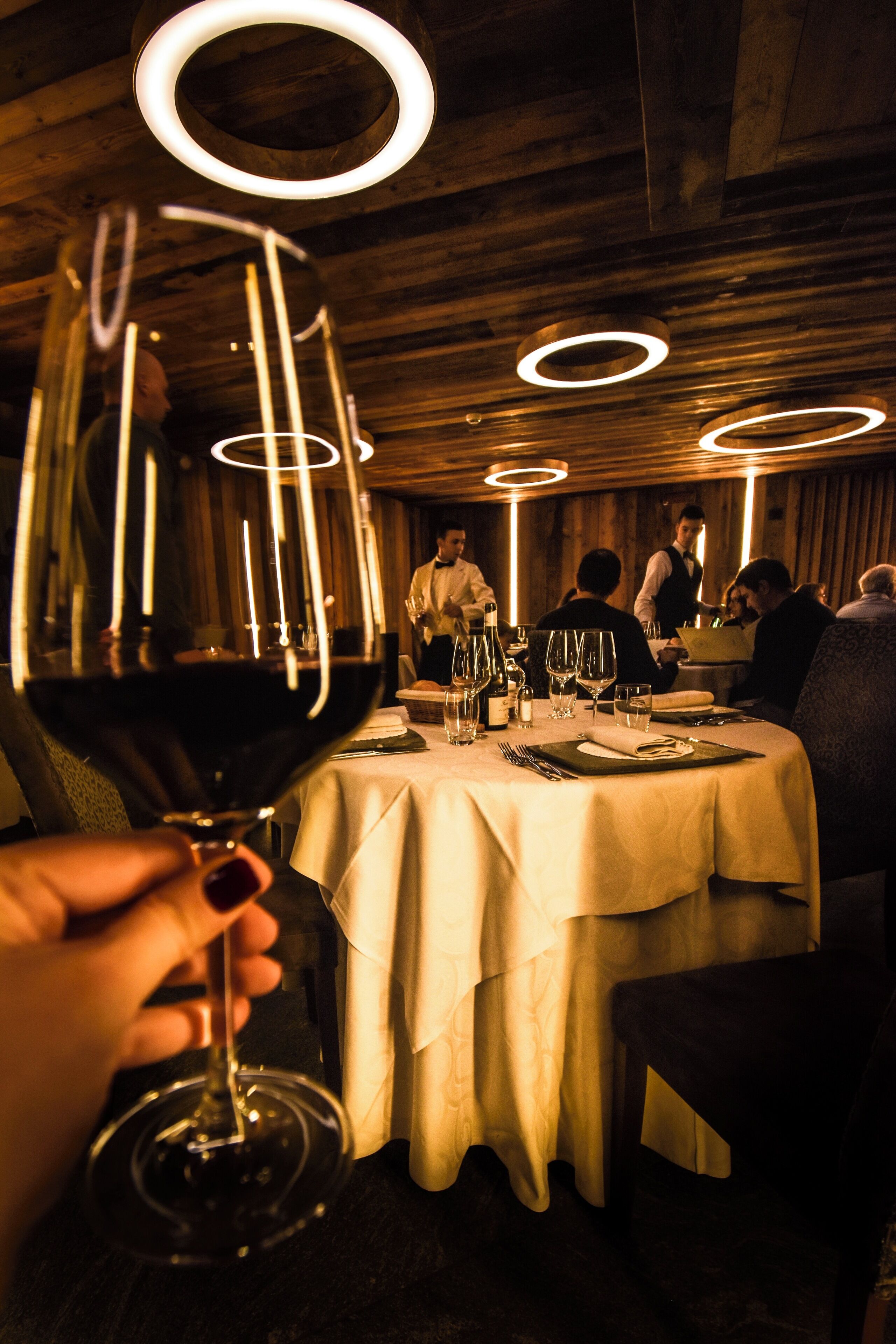 A restaurant with round ceiling lights, a table set for dining, and a person holding a wine glass.