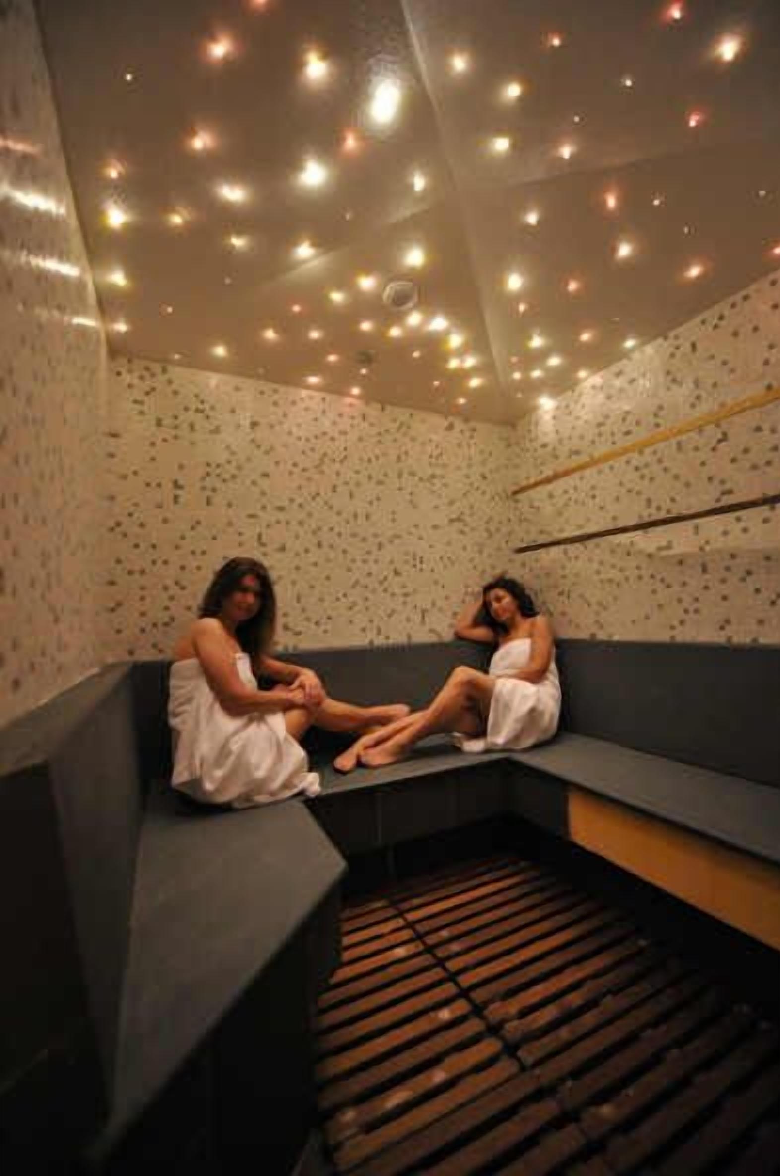A spa room with two people sitting on a bench, a wooden slatted floor, and a ceiling with numerous lights.