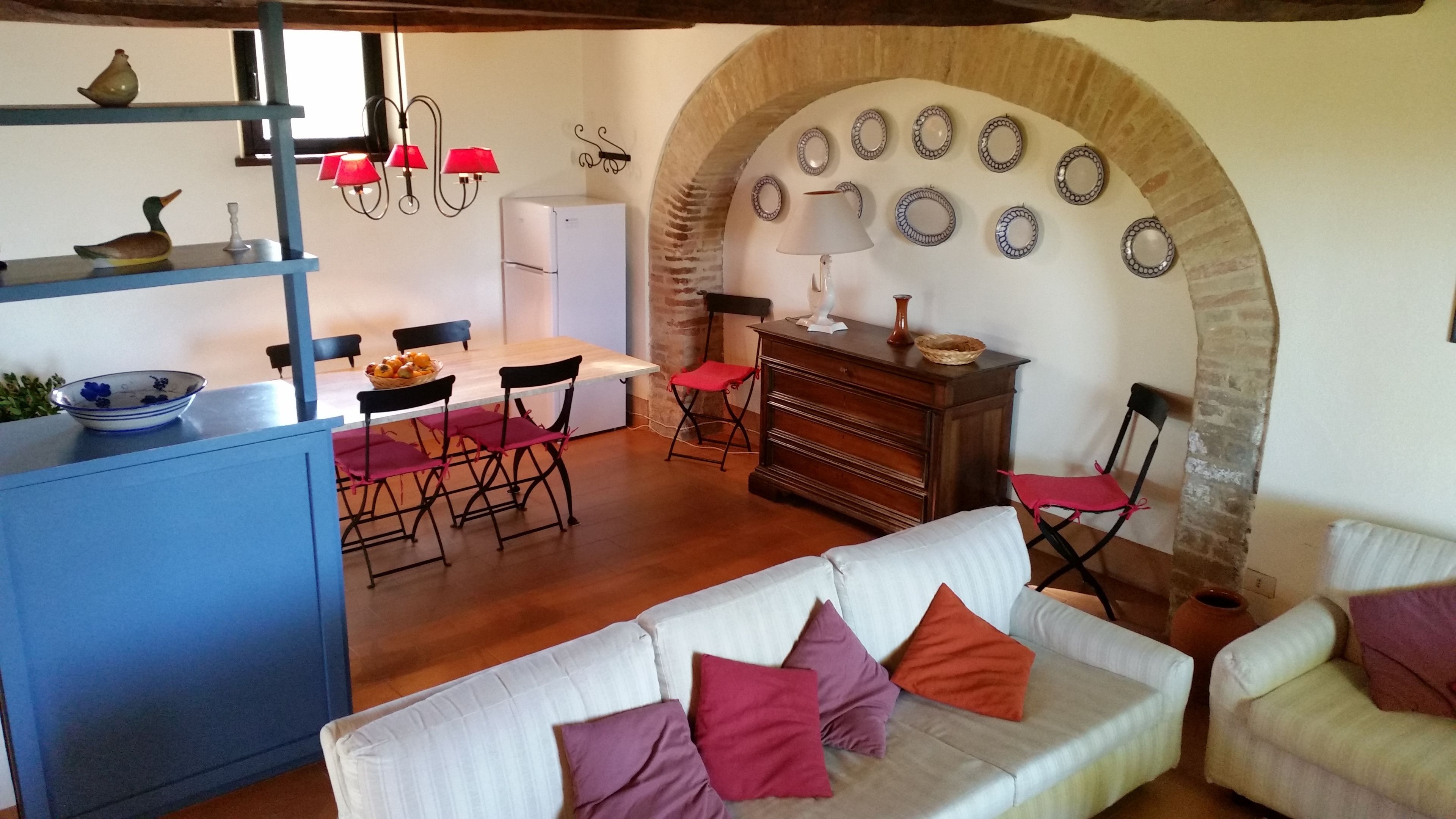 A cozy living room with a white sofa, red cushions, a dining area with a wooden table and chairs, and a kitchen with a blue cabinet and decorative items.