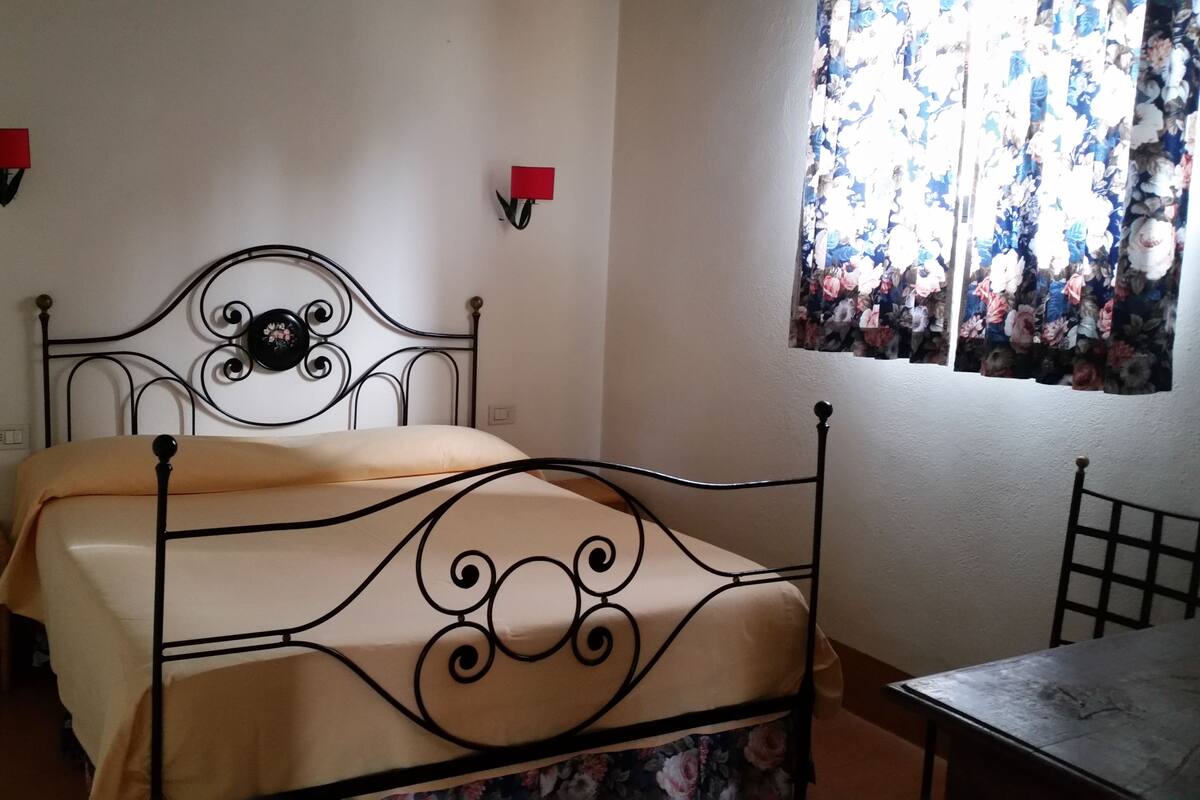 Un lit simple avec un cadre en métal et une couvre-lit beige, une petite table en bois et une fenêtre ornée de rideaux à motifs floraux.