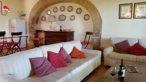 A living room with a white sofa, red and purple cushions, a small table with wine glasses and a bottle, and a brick wall with decorative plates. - Agriturismo Poggio alle Vigne (Torgiano)