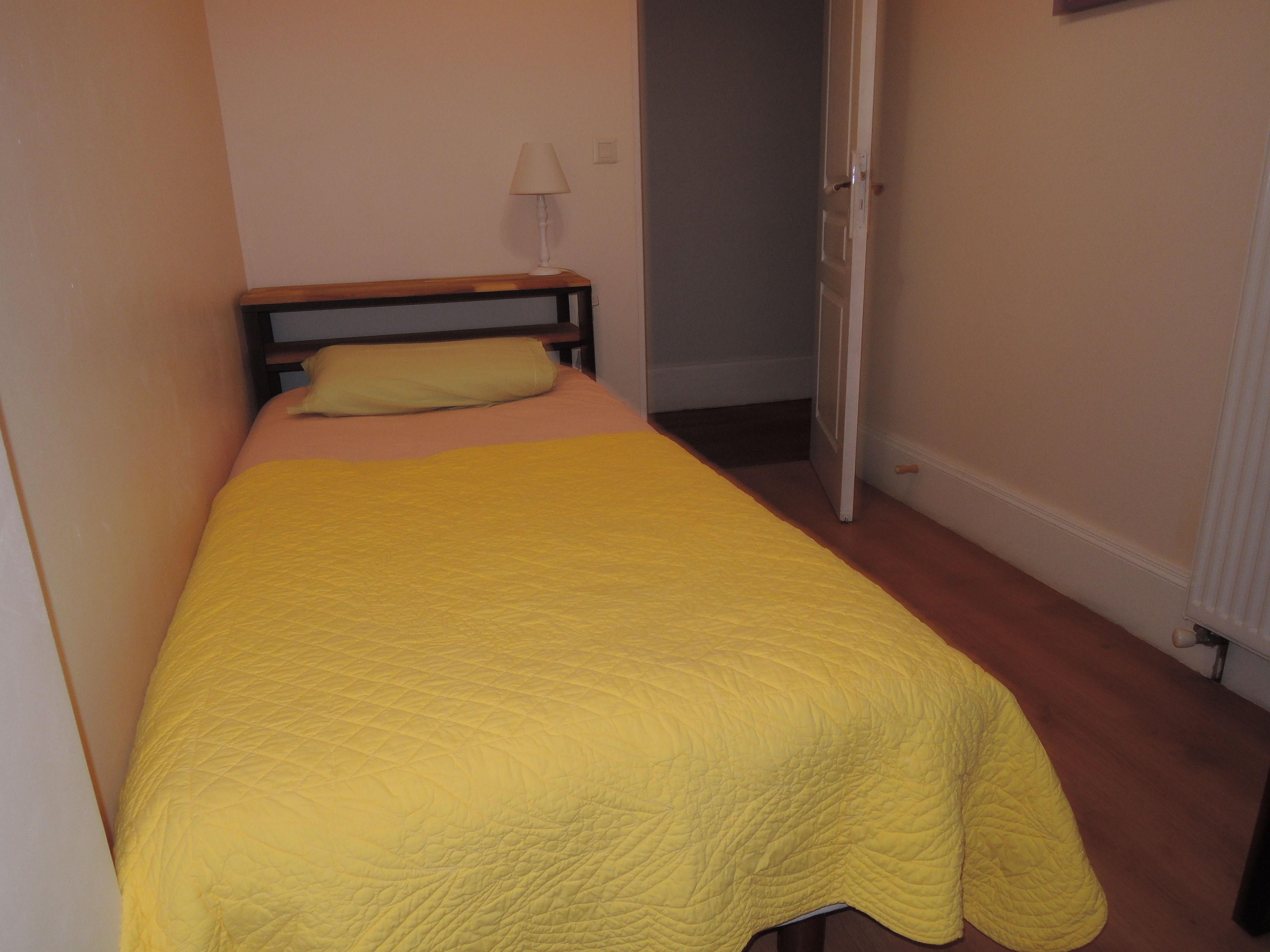 Una habitación pequeña con cama individual, cubrecama amarillo, cabecera de madera y puerta que da a otra habitación.