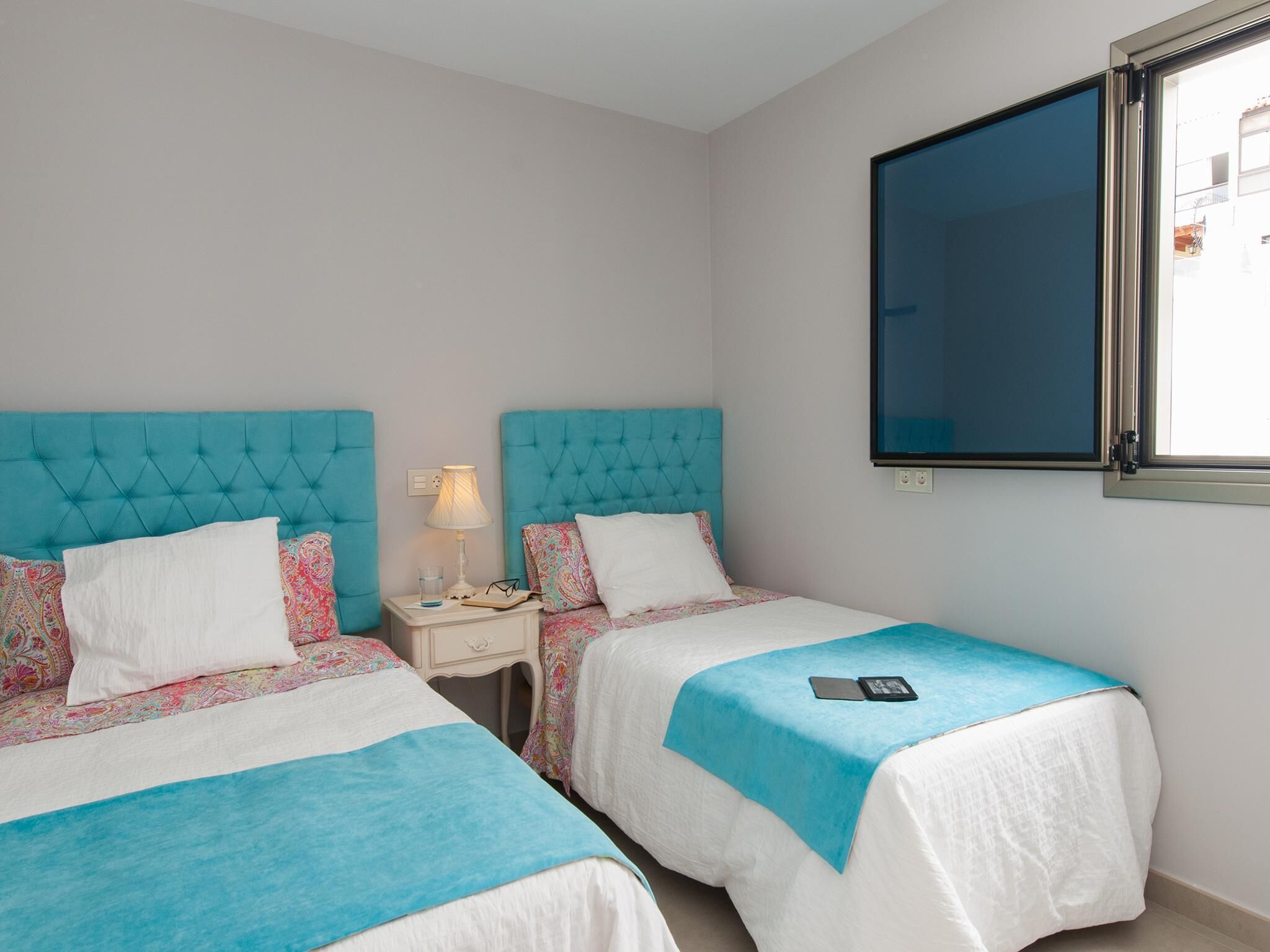 A bedroom with two beds, each with a blue headboard and white bedding. There is a window on the right wall, and a nightstand with a lamp and a glass of water between the beds.