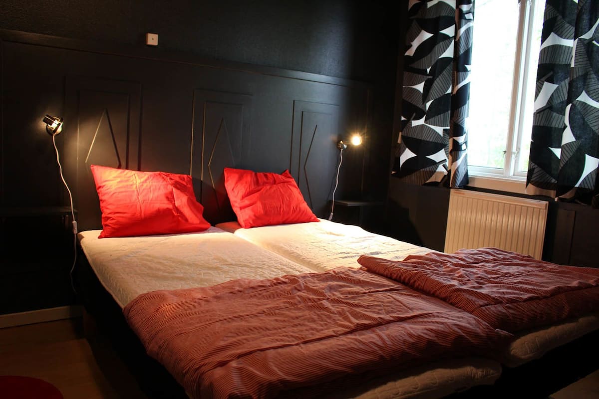Une chambre à coucher avec un lit, deux oreillers rouges, un couvre-lit blanc et une couverture rouge. On y trouve deux lampes de chevet et une fenêtre avec des rideaux à motifs.
