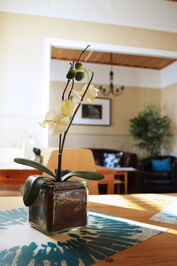 Un florero de vidrio con una planta sobre una mesa de madera en una habitación con una mesa de comedor y sillas, un sofá y otra planta al fondo.