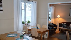 Un coin repas avec une table en bois, des chaises et des plantes en pot. On aperçoit un canapé et une lampe au fond.