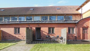 A two-story brick building with a tiled roof, a covered patio with a table and chairs, and a small garden area.